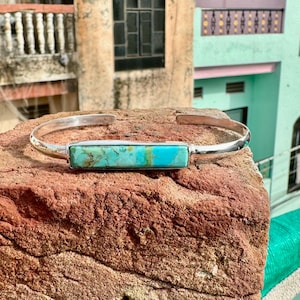May include: A silver cuff bracelet featuring a rectangular turquoise stone. The stone displays shades of blue and green. The bracelet is presented on a rough, reddish-brown brick surface.