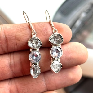 Peut inclure: Boucles d'oreilles pendantes en argent, chacune avec trois pierres précieuses : une pierre grise de forme irrégulière en haut, une pierre ronde et transparente au milieu et une pierre transparente en forme de diamant en bas. Les boucles d'oreilles sont tenues par une main.