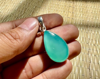 May include: A teardrop-shaped pendant with a vibrant, translucent teal stone. The stone is set in a silver-colored metal. The pendant is held in a hand against a textured, light brown background.