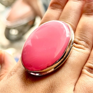Peut inclure: Une bague ovale avec une grosse pierre rose vif sertie dans un anneau argenté. La pierre polie a une finition brillante, reflétant la lumière. Ce bijou est un accessoire audacieux.
