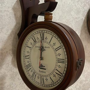 May include: A wooden wall clock with a round face and Roman numerals. The clock face is cream-colored with black numerals and hands. The clock is mounted on a dark brown wooden bracket. The clock face reads "Jeffers & Smith London".