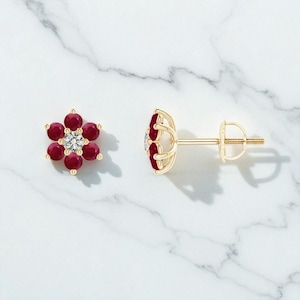 May include: A pair of gold-coloured stud earrings in a flower design. Each earring has six round, deep red gemstones encircling a central, clear gemstone. The earrings are presented on a white marble surface.