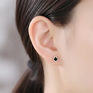May include: Close-up of a person's ear with a gold stud earring. The earring features a black four-leaf clover design with a gold border and small white accents. The earring is a small, delicate piece of jewelry.
