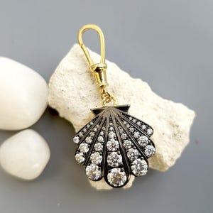 May include: A seashell-shaped charm with a gold-coloured clasp and a dark grey body. The charm is encrusted with numerous clear, sparkling gemstones. The charm is resting on a piece of white stone, with two white stones nearby.