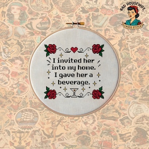 May include: A circular wooden embroidery hoop containing white fabric with a cross-stitch design. The text reads, I invited her into my home. I gave her a beverage. The design includes four red roses, a small red heart, and a martini glass icon.