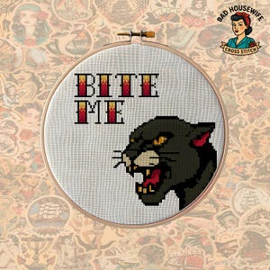 Puede incluir: Bordado de punto de cruz en un aro de madera. El diseño presenta las palabras "BITE ME" en letras de bloque rojas y amarillas, y una pantera negra con ojos amarillos y boca roja. El fondo es blanco.