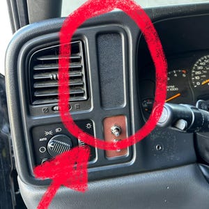May include: Close-up of a black car dashboard with various controls. Features include air vents, headlight controls, and a toggle switch on a brown panel. The dashboard is made of black plastic.