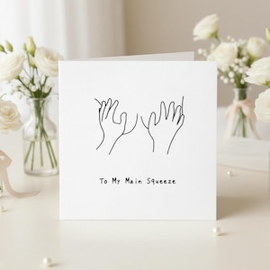 May include: A white greeting card with a minimalist line drawing of two hands reaching out to each other. The text "To My Main Squeeze" is printed below the illustration. The card is surrounded by white roses in glass vases.