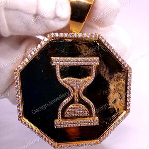 May include: A gold-colored pendant with an octagonal shape and a black center. The pendant features an hourglass design encrusted with small, clear gemstones. The outer edge is also lined with gemstones.