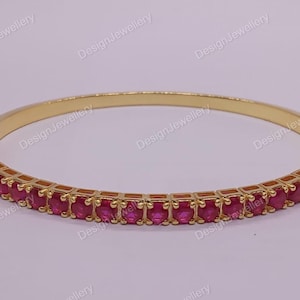 May include: A gold-coloured bangle bracelet featuring a line of vivid, oval-cut, red gemstones. The bracelet has a hinged clasp and is set against a light background. The text "DesignJewellery" is visible.