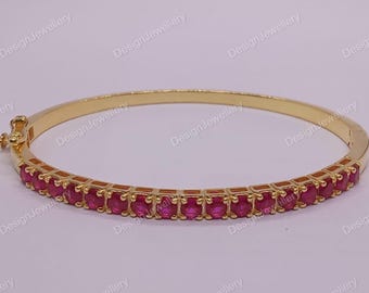 Könnte beinhalten: Ein goldfarbenes Armreif-Armband mit einer Reihe von leuchtenden, oval geschliffenen, roten Edelsteinen. Das Armband hat einen Klappverschluss und ist vor einem hellen Hintergrund platziert. Der Text "DesignJewellery" ist sichtbar.