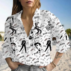 May include: White button-down shirt featuring a repeating pattern of Elvis Presley images, musical notes, guitars, and the word "ELVIS". The shirt has long sleeves and a collared neckline. The design includes various poses of Elvis.
