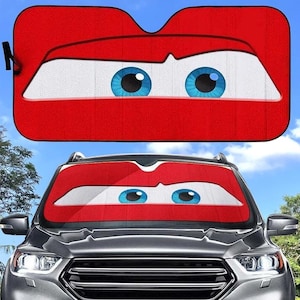 Op de afbeelding: Rode en witte auto zonnescherm met een cartoon gezicht, met blauwe ogen en een rode mond. Het zonnescherm is geïnstalleerd op een autoruit en biedt bescherming tegen de zon.
