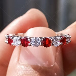 May include: A silver ring with alternating red and clear gemstones. The ring is held between fingers, showcasing the vibrant colors of the gemstones. The ring is a band style, perfect for everyday wear.