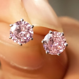 May include: A pair of stud earrings featuring round, light pink gemstones set in a silver metal. The earrings are held up close to the camera, showcasing the sparkle and color of the stones. The earrings are a classic jewelry item.