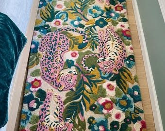 May include: A rectangular tufted rug featuring a cream-colored background with a vibrant pattern of pink leopards with dark spots, interspersed with large teal, yellow, and pink flowers and green tropical leaves across the entire surface.