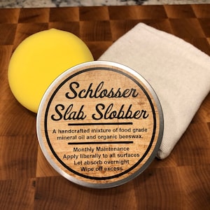 May include: A kit for wood surface care, including a yellow sponge, a white cloth, and a metal tin labeled "Schlosser Slab Slobber." The tin contains a handcrafted mixture of mineral oil and beeswax for monthly maintenance.