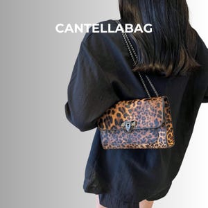 Puede incluir: Un bolso pequeño y rectangular con estampado de leopardo. El bolso tiene un cierre plateado y una correa de cadena. El texto "CANTELLABAG" está impreso en blanco en la parte superior.