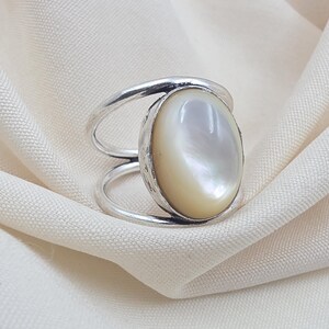 Può includere: Un anello in argento con un grande cabochon ovale in madreperla iridescente incastonato in una montatura in metallo lucido. La fascia è composta da due fili separati, lisci e color argento, che convergono ai lati per sostenere la pietra.