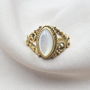 May include: A gold-toned ring featuring a central marquise-shaped white mother-of-pearl cabochon. The metal band displays intricate scrollwork, twisted rope detailing around the stone setting, and small spherical accents along the sides.