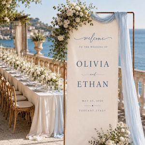 May include: A tall, rectangular wedding welcome sign on a copper frame, featuring cream fabric with blue text reading: Welcome to the wedding of Olivia and Ethan, May 24, 2026, Tuscany, Italy. It is draped with light blue fabric and white floral arrangements.