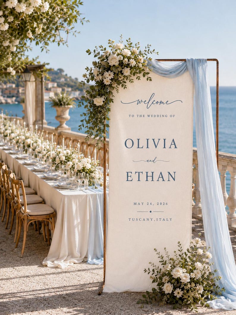 May include: A tall, rectangular wedding welcome sign on a copper frame, featuring cream fabric with blue text reading: Welcome to the wedding of Olivia and Ethan, May 24, 2026, Tuscany, Italy. It is draped with light blue fabric and white floral arrangements.