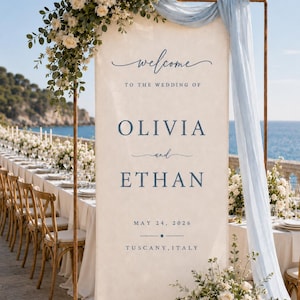 May include: A tall, rectangular fabric wedding welcome sign in cream with blue text reading "welcome TO THE WEDDING OF OLIVIA and ETHAN MAY 24, 2026 TUSCANY, ITALY". The sign hangs from a copper frame draped with light blue fabric and white floral arrangements.