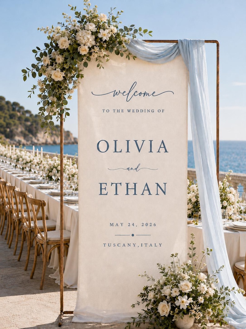 May include: A tall, rectangular fabric wedding welcome sign in cream with blue text reading "welcome TO THE WEDDING OF OLIVIA and ETHAN MAY 24, 2026 TUSCANY, ITALY". The sign hangs from a copper frame draped with light blue fabric and white floral arrangements.