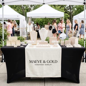Può includere: Un tavolo nero coperto da un runner color crema con il testo MAEVE & GOLD HANDMADE JEWELRY. Il tavolo espone vari gioielli su supporti beige, tra cui collane su busti, braccialetti su barre e piccole scatole per anelli.