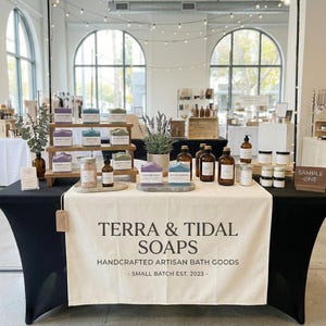 May include: A display of handcrafted artisan bath goods, including soaps, oils, and bath salts, arranged on a table with a white tablecloth. The table features the brand name "TERRA & TIDAL SOAPS" and the text "HANDCRAFTED ARTISAN BATH GOODS - SMALL BATCH EST. 2023-".