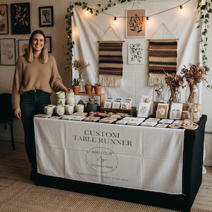 May include: A craft fair booth featuring a white table runner with the text "CUSTOM TABLE RUNNER" and "ADD YOUR LOGO OR FAMILY NAME". The table displays handmade mugs, cards, and woven wall hangings. A woman stands next to the table.