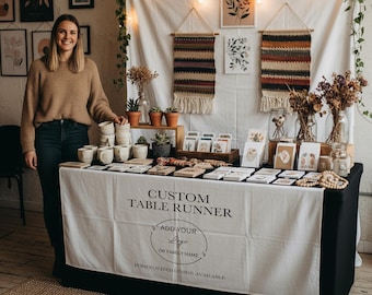 Puede incluir: Un puesto de feria artesanal con un mantel blanco con el texto "CUSTOM TABLE RUNNER" y "ADD YOUR LOGO OR FAMILY NAME". La mesa exhibe tazas hechas a mano, tarjetas y tapices de pared tejidos. Una mujer está de pie junto a la mesa.