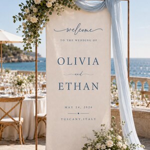 May include: A tall, rectangular cream-coloured fabric wedding welcome sign suspended on a copper-toned metal frame. The sign features dark blue text reading "welcome TO THE WEDDING OF OLIVIA and ETHAN MAY 24, 2026 TUSCANY, ITALY" with floral arrangements.