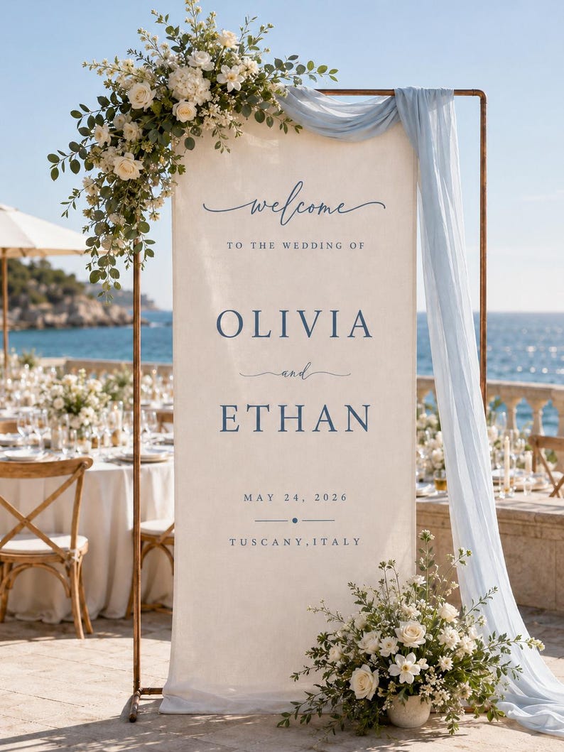 May include: A tall, rectangular cream-coloured fabric wedding welcome sign suspended on a copper-toned metal frame. The sign features dark blue text reading "welcome TO THE WEDDING OF OLIVIA and ETHAN MAY 24, 2026 TUSCANY, ITALY" with floral arrangements.