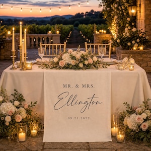 Puede incluir: Un camino de mesa de tela color crema con el texto impreso MR. & MRS. Ellington y la fecha 09.21.2027. El camino está colocado sobre una mesa decorada con un centro de mesa floral, velas y cristalería en un entorno exterior al atardecer.