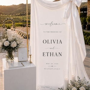 May include: A white fabric wedding welcome sign draped over a gold metal frame, featuring a large satin bow at the top right. The text reads: welcome TO THE WEDDING OF OLIVIA and ETHAN MAY 24, 2026 TUSCANY, ITALY in black serif and script fonts.