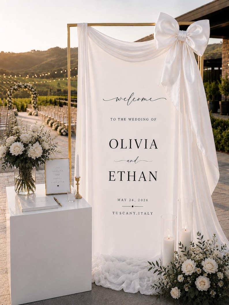 May include: A white fabric wedding welcome sign draped over a gold metal frame, featuring a large satin bow at the top right. The text reads: welcome TO THE WEDDING OF OLIVIA and ETHAN MAY 24, 2026 TUSCANY, ITALY in black serif and script fonts.