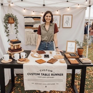Puede incluir: Una mujer está detrás de una mesa que exhibe artículos de cuero y tazas. La mesa tiene un camino de mesa personalizado. Los productos incluyen carteras, posavasos y bandejas de madera. El fondo presenta un tapiz tejido y luces de cadena.