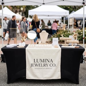 Può includere: Un tavolo espositivo nero per Lumina Jewelry Co. con un runner color crema riportante il nome del marchio, vari espositori per gioielli, uno specchio rotondo, busti per collane e diversi vassoi contenenti anelli e bracciali in un mercato.