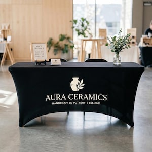 May include: A black table cover with the white text "AURA CERAMICS HANDCRAFTED POTTERY | Est. 2021" and a logo of a vase and leaves. A small sign displays "PRICE CERAMICS" and prices for mugs, bowls, and vases. A vase of greenery sits on the table.