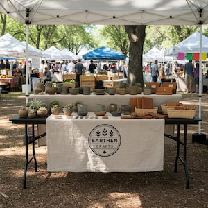May include: A stall at an outdoor market showcases handmade pottery, such as mugs, bowls, and succulent planters. Leather items and woven baskets are also on display on a table covered with a linen cloth. The vendor's logo, "Earthen Crafts," is visible.
