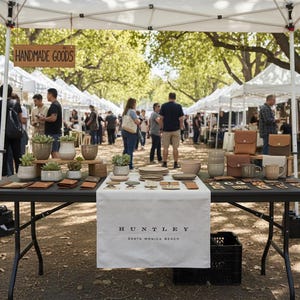 May include: An outdoor market scene with various handmade goods displayed on tables. The tables showcase items like leather wallets, ceramic bowls, and potted succulents. A sign reads "HANDMADE GOODS". The banner reads "HUNTLEY SANTA MONICA BEACH".
