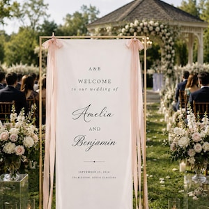 May include: A rectangular white wedding welcome sign held by a gold metal frame, decorated with light pink ribbons at the top corners. The text reads: A & B, WELCOME to our wedding of, Amelia AND Benjamin, SEPTEMBER 29, 2026, CHARLESTON, SOUTH CAROLINA.