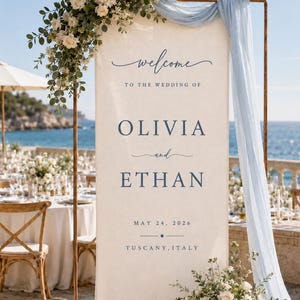 Könnte beinhalten: Ein hohes, rechteckiges Hochzeits-Willkommensschild aus cremefarbenem Stoff, das an einem kupferfarbenen Metallrahmen hängt. Das Schild zeigt dunkelblauen Text mit der Aufschrift "welcome TO THE WEDDING OF OLIVIA and ETHAN MAY 24, 2026 TUSCANY, ITALY" und Blumenschmuck.