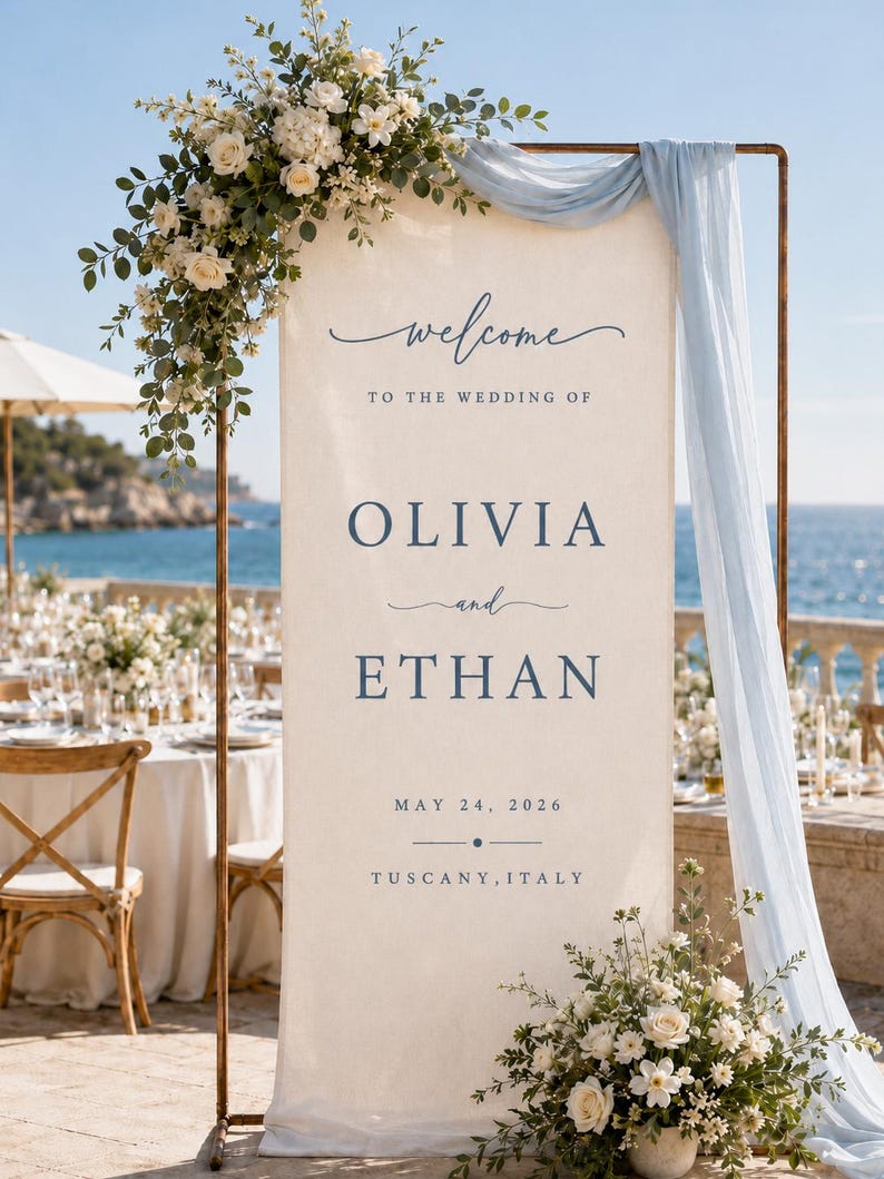 May include: A tall, rectangular cream-coloured fabric wedding welcome sign suspended on a copper-toned metal frame. The sign features dark blue text reading "welcome TO THE WEDDING OF OLIVIA and ETHAN MAY 24, 2026 TUSCANY, ITALY" with floral arrangements.