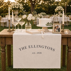 Puede incluir: Un camino de mesa de tela color crema sobre una mesa de madera, con el texto impreso en negro THE ELLINGTONS est. 2026. La mesa está decorada con un pastel blanco en un soporte, un jarrón con flores blancas, copas de cristal y una foto.