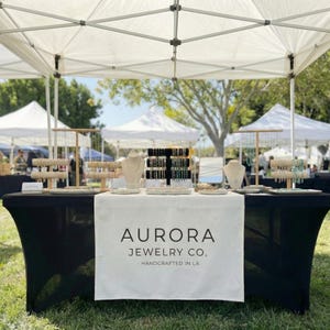 Puede incluir: Un puesto de joyería en un mercado al aire libre bajo una carpa blanca. La mesa está cubierta con un mantel negro y un camino de mesa blanco con el texto "AURORA JEWELRY CO. - HANDCRAFTED IN LA." Se exhiben varios collares, pulseras y aretes.