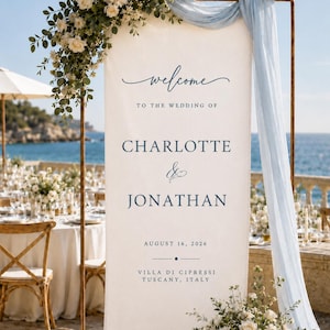 May include: A tall, rectangular white wedding welcome sign suspended on a copper-coloured metal frame, featuring dark blue text reading "welcome TO THE WEDDING OF CHARLOTTE & JONATHAN AUGUST 16, 2026 VILLA DI CIPRESSI TUSCANY, ITALY" with floral arrangements.