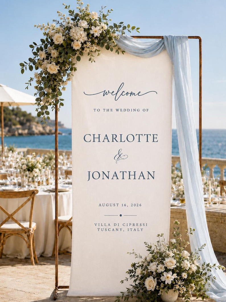 May include: A tall, rectangular white wedding welcome sign suspended on a copper-coloured metal frame, featuring dark blue text reading "welcome TO THE WEDDING OF CHARLOTTE & JONATHAN AUGUST 16, 2026 VILLA DI CIPRESSI TUSCANY, ITALY" with floral arrangements.