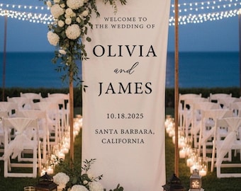 May include: A wedding ceremony setup with a banner that reads "Welcome to the Wedding of Olivia and James" with the date 10.18.2025 and location Santa Barbara, California. The banner is decorated with white flowers and greenery. White chairs are set up for guests.